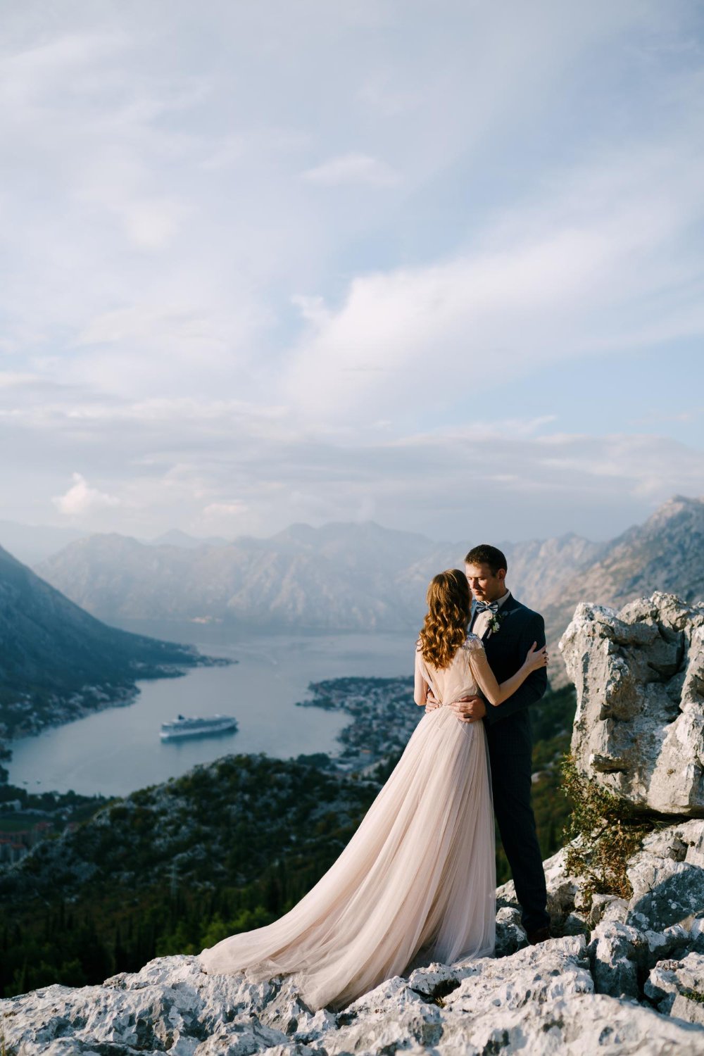 Groom hugs bride by the waist on a rocky mountain overlooking the bay