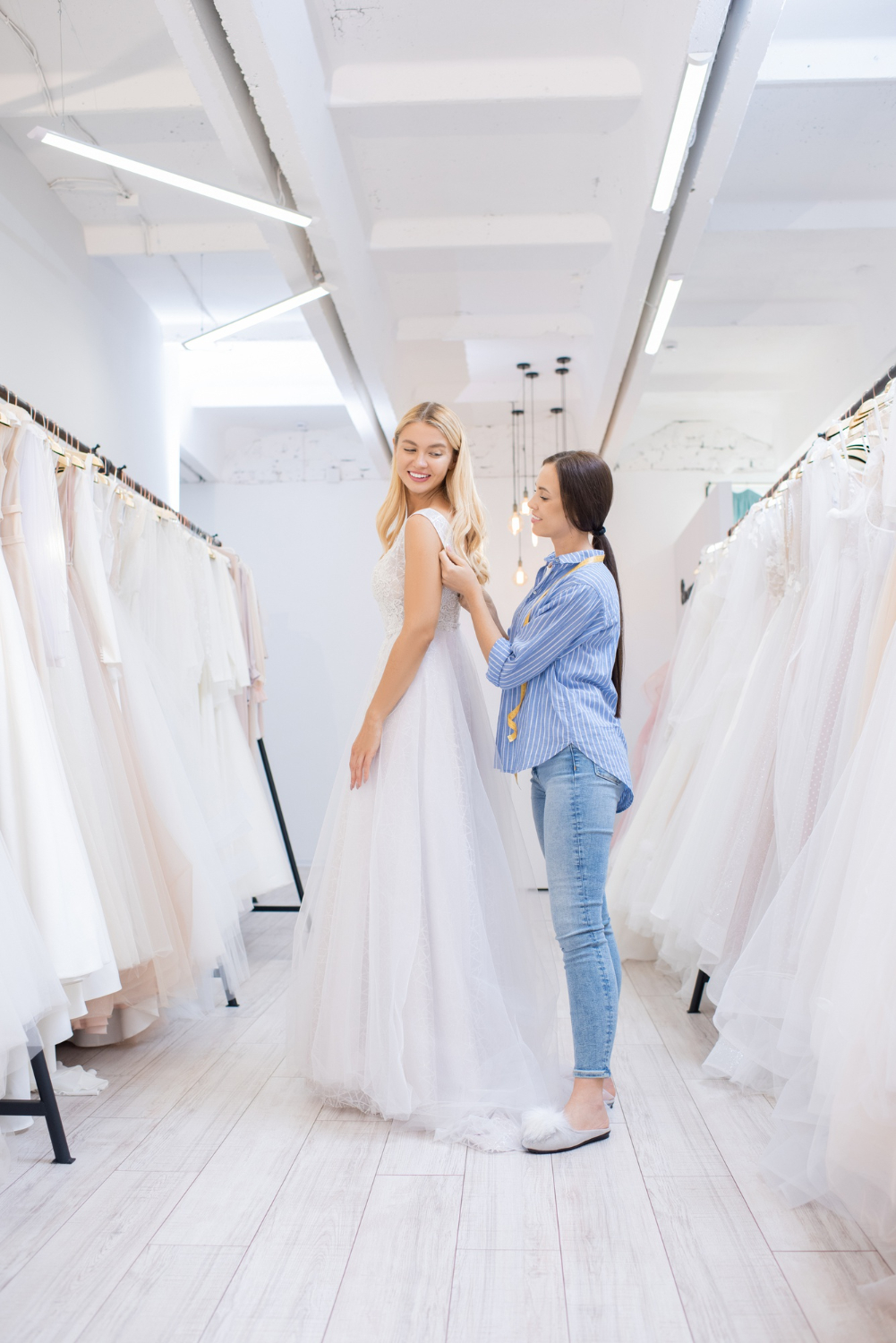 smiling brunette wedding dress tailor assisting bride to wear dress between rows of beautiful gowns