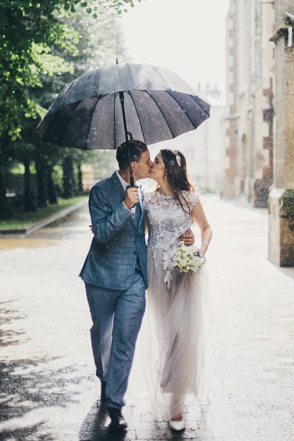stylish bride and groom walking under umbrella and kissing on background of old church in rain